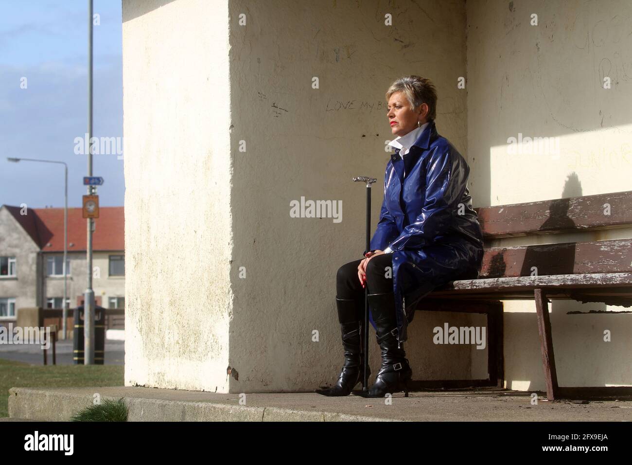 White caucasian middle aged woman with cropped spikey dyed hair wearing blue PVC raincoat sat on bench at river harbour location looking pensive, puzzled, lost Stock Photo