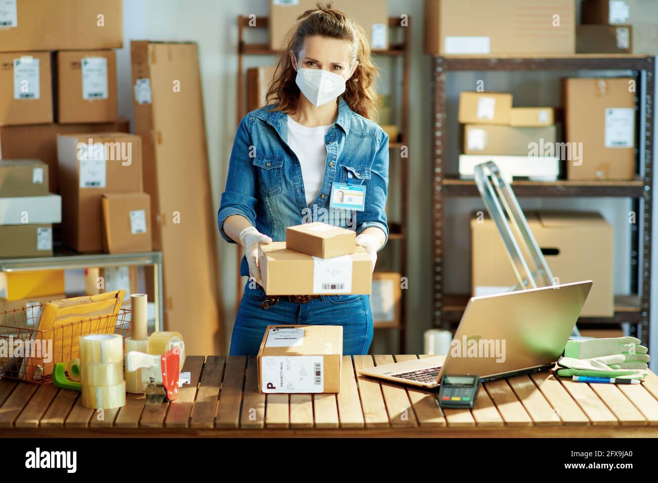 Delivery business. Portrait of modern female in jeans with laptop and ffp2 mask giving parcel in the office. Stock Photo