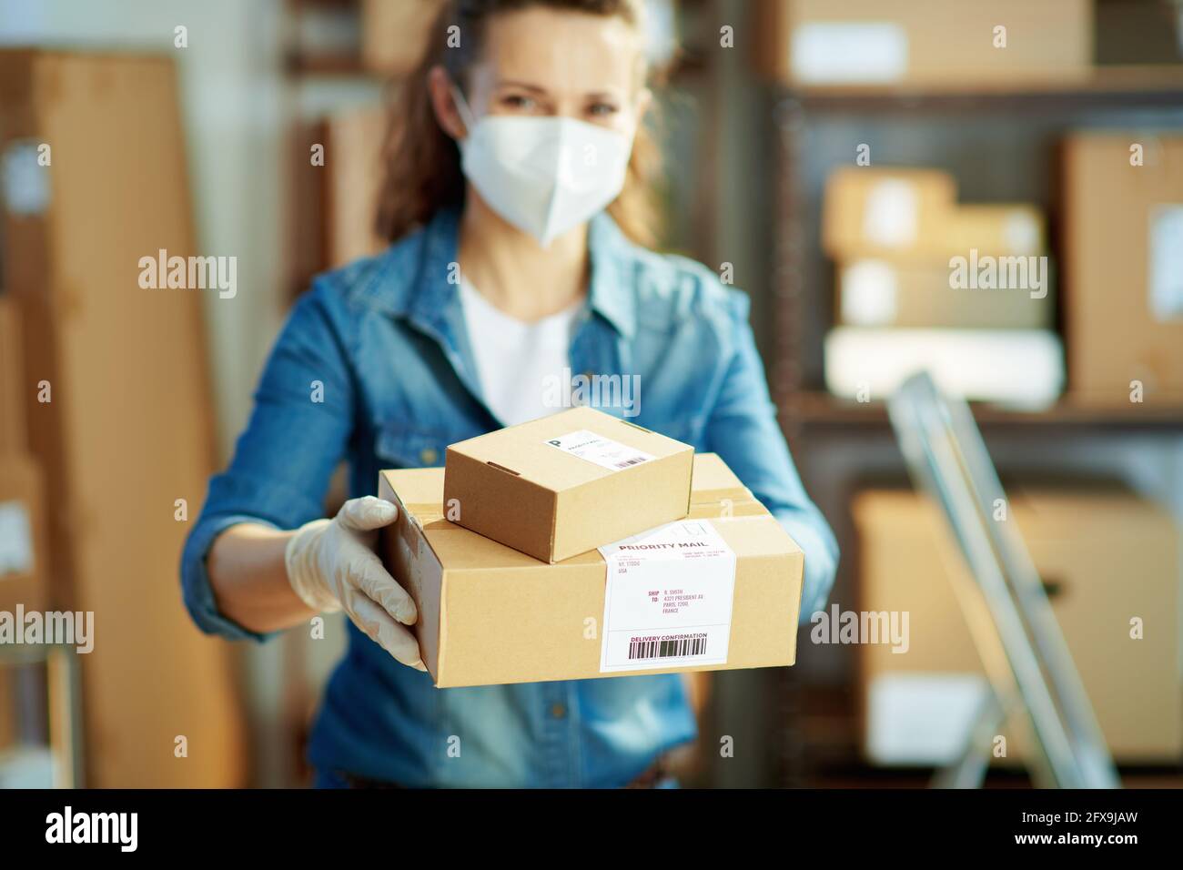 Delivery business. Closeup on woman in jeans with ffp2 mask giving parcel in the warehouse. Stock Photo