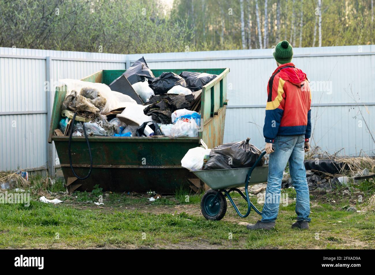 Young man in a jacket throws the garbage brought from his homestead into a common village dumpster in cold spring evening. Stock Photo
