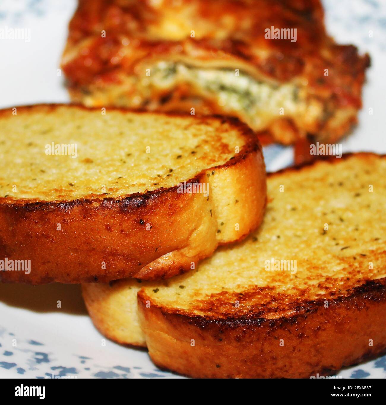 Close-up of two slices of garlic bread with a slice of homemade lasagna out of focus in background. Stock Photo