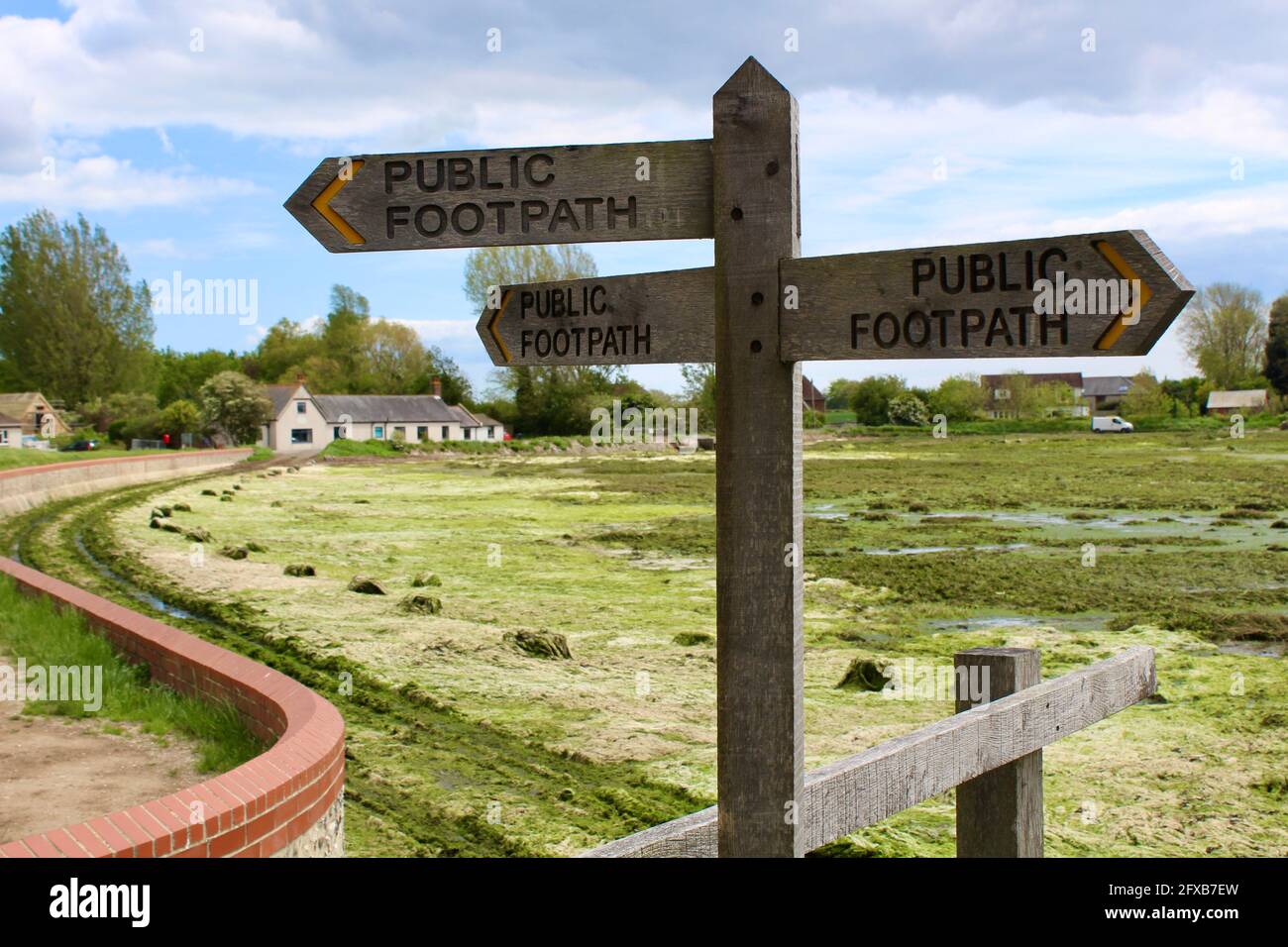 Public footpath sign at Bosham harbour with the old school house in the background. High tide left much seaweed on the road. Car tracks visible. Stock Photo