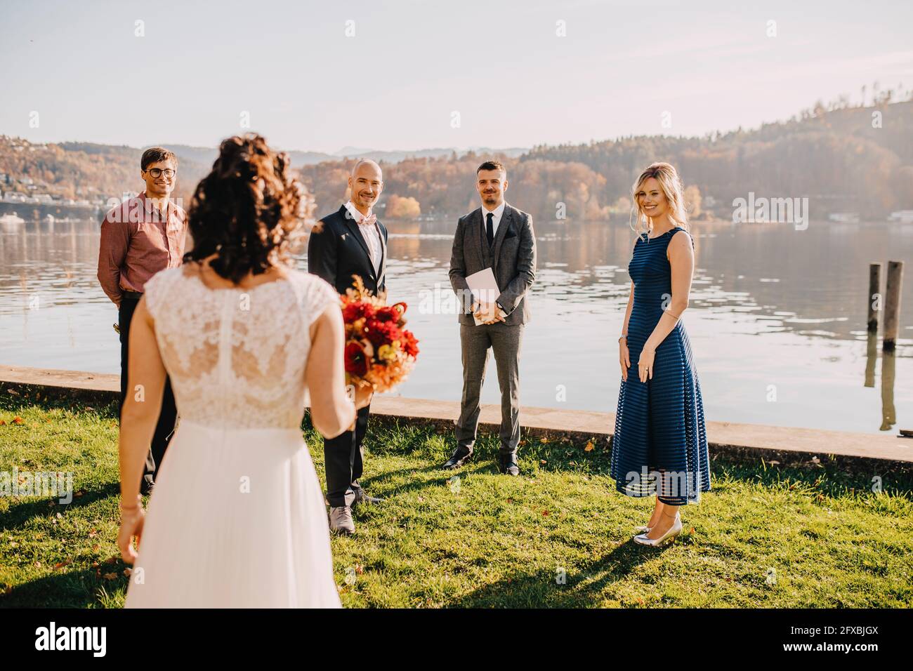 Groom standing with wedding guests looking at bride in front of lake on wedding ceremony Stock Photo