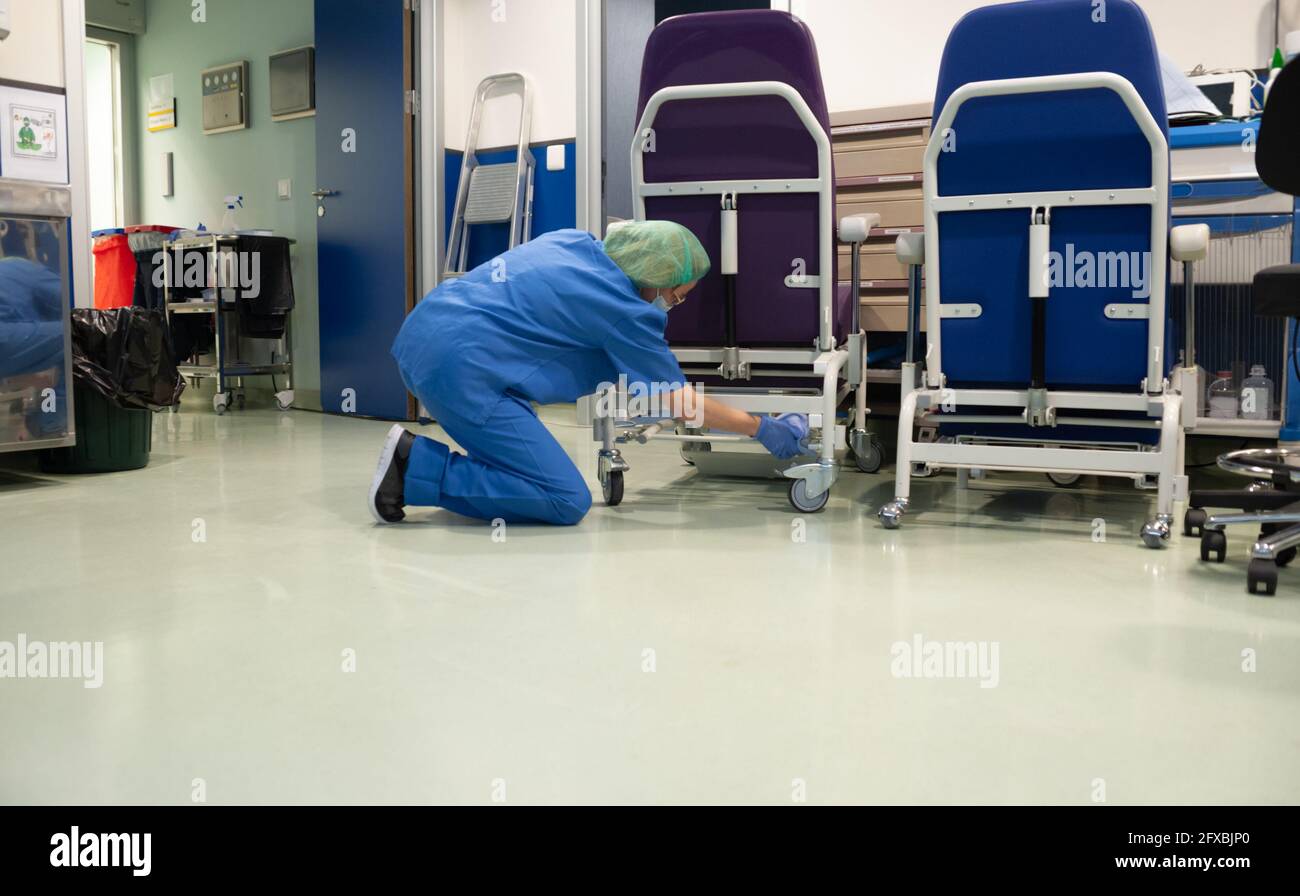 Woman from the cleaning service of a hospital crouching cleaning the wheels of a patient chair in the operating room beforehand. Sanitary concept Stock Photo