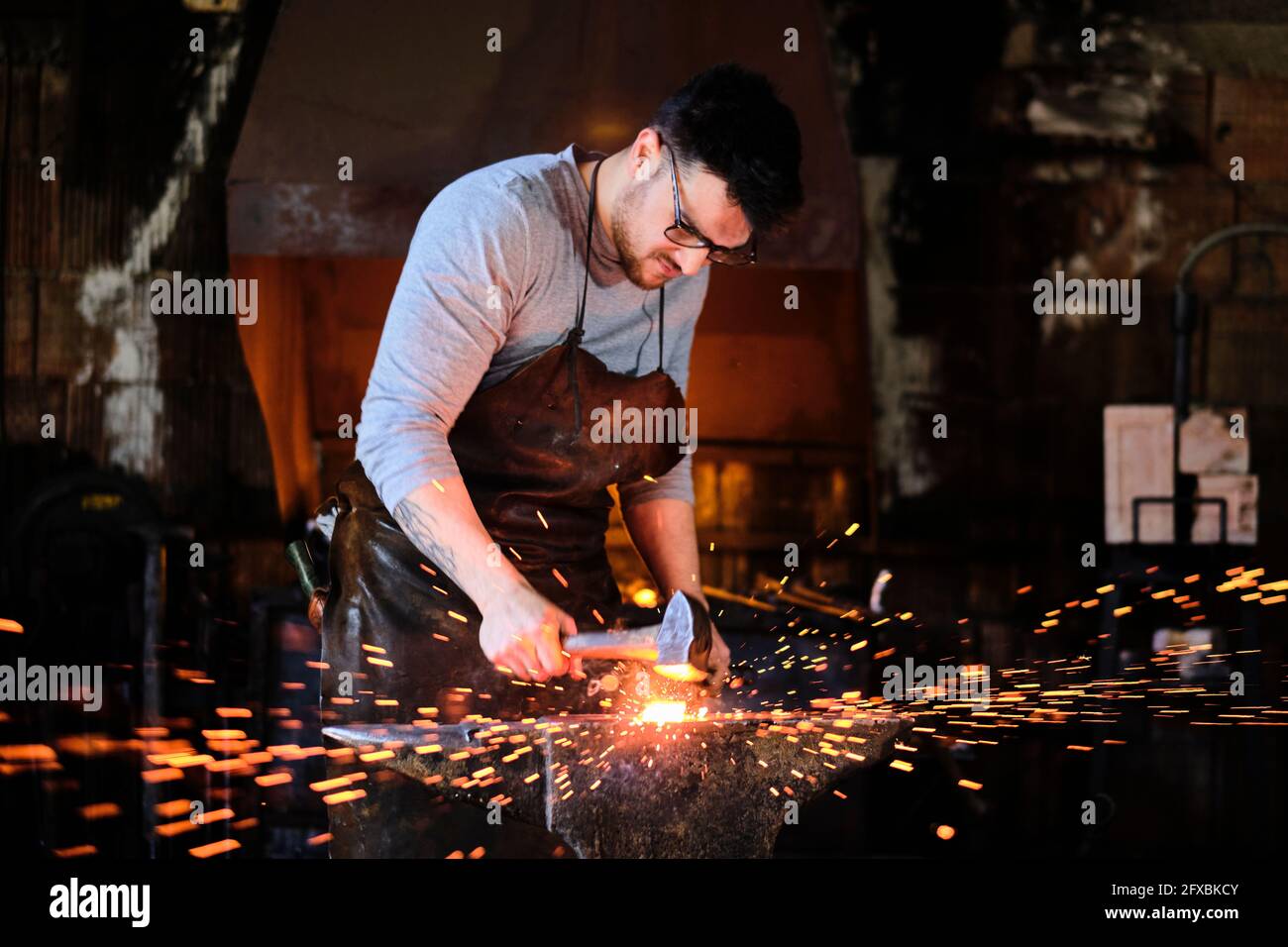 Young male blacksmith wearing eyeglasses forging with hammer at workshop Stock Photo