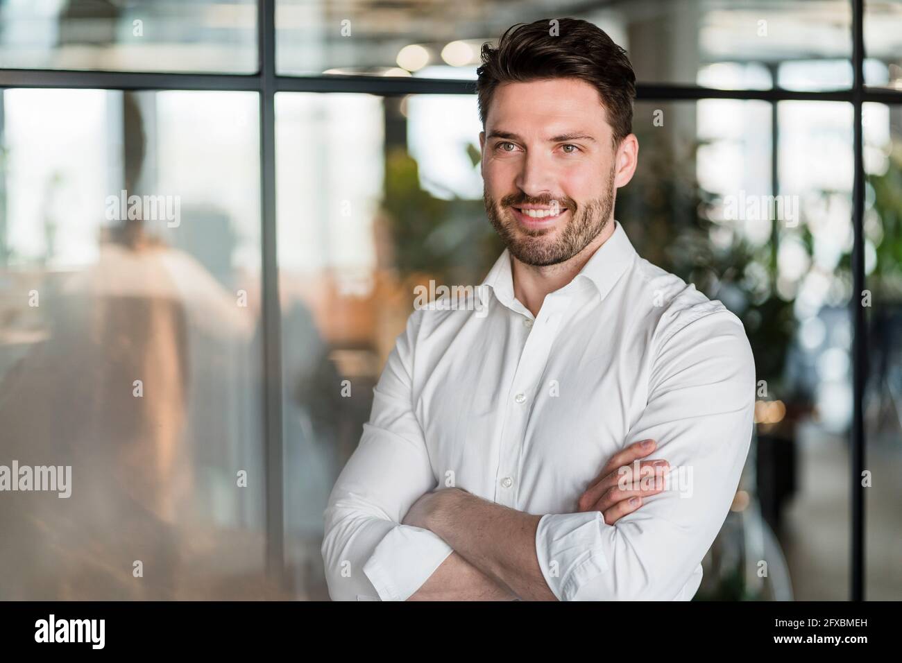 Smiling male professional with arms crossed standing by glass in office ...