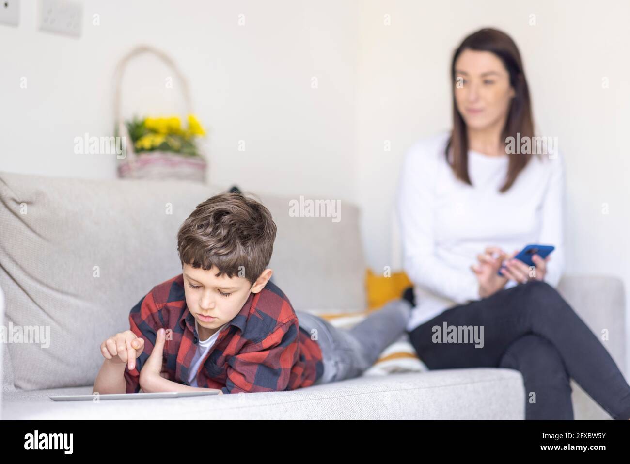 Boy using digital tablet while lying on sofa with mother in background ...