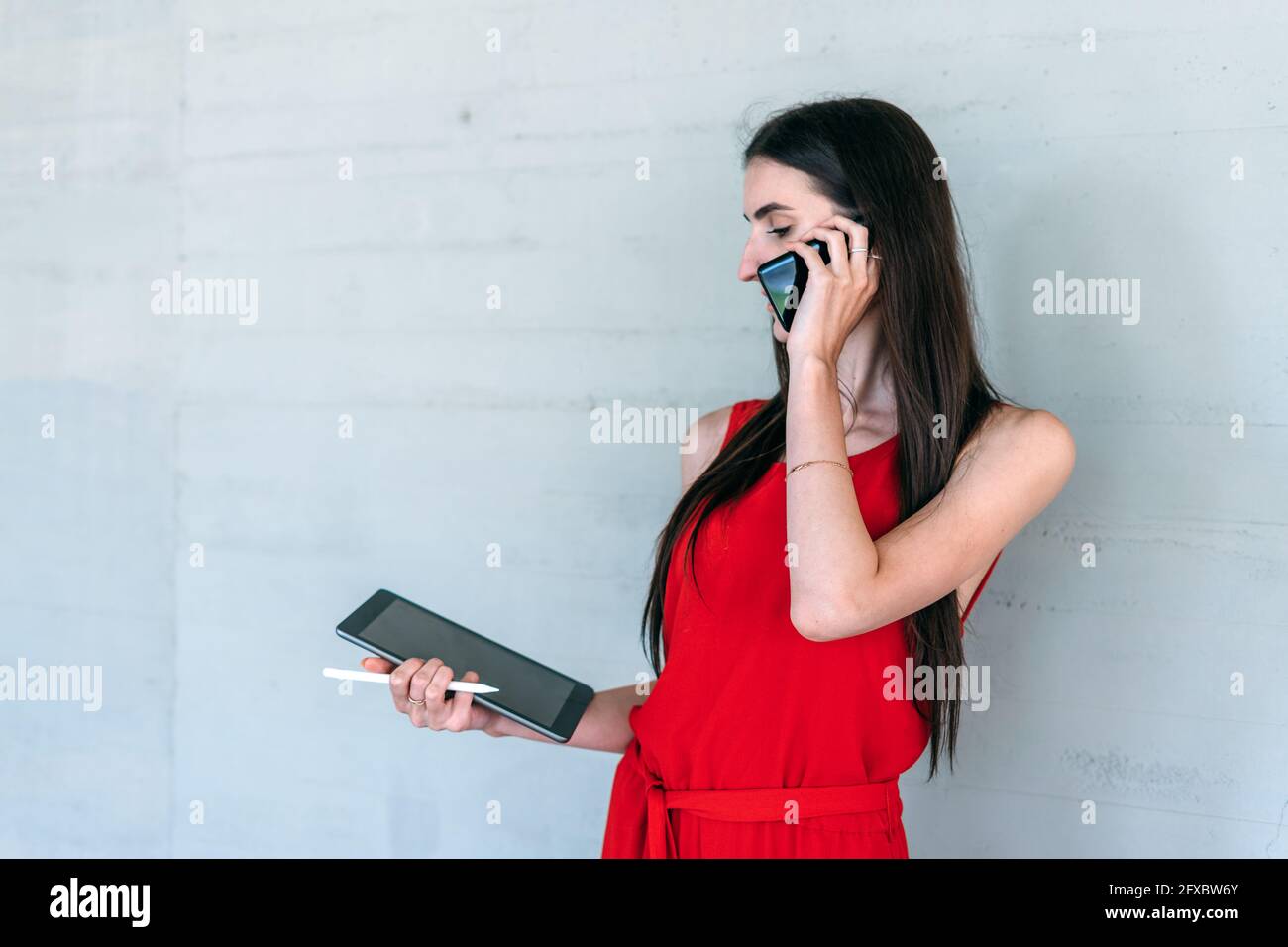 Young business woman in red dress, holding a tablet while receiving a phone call with her cell phone looking at the digital tablet. Stock Photo