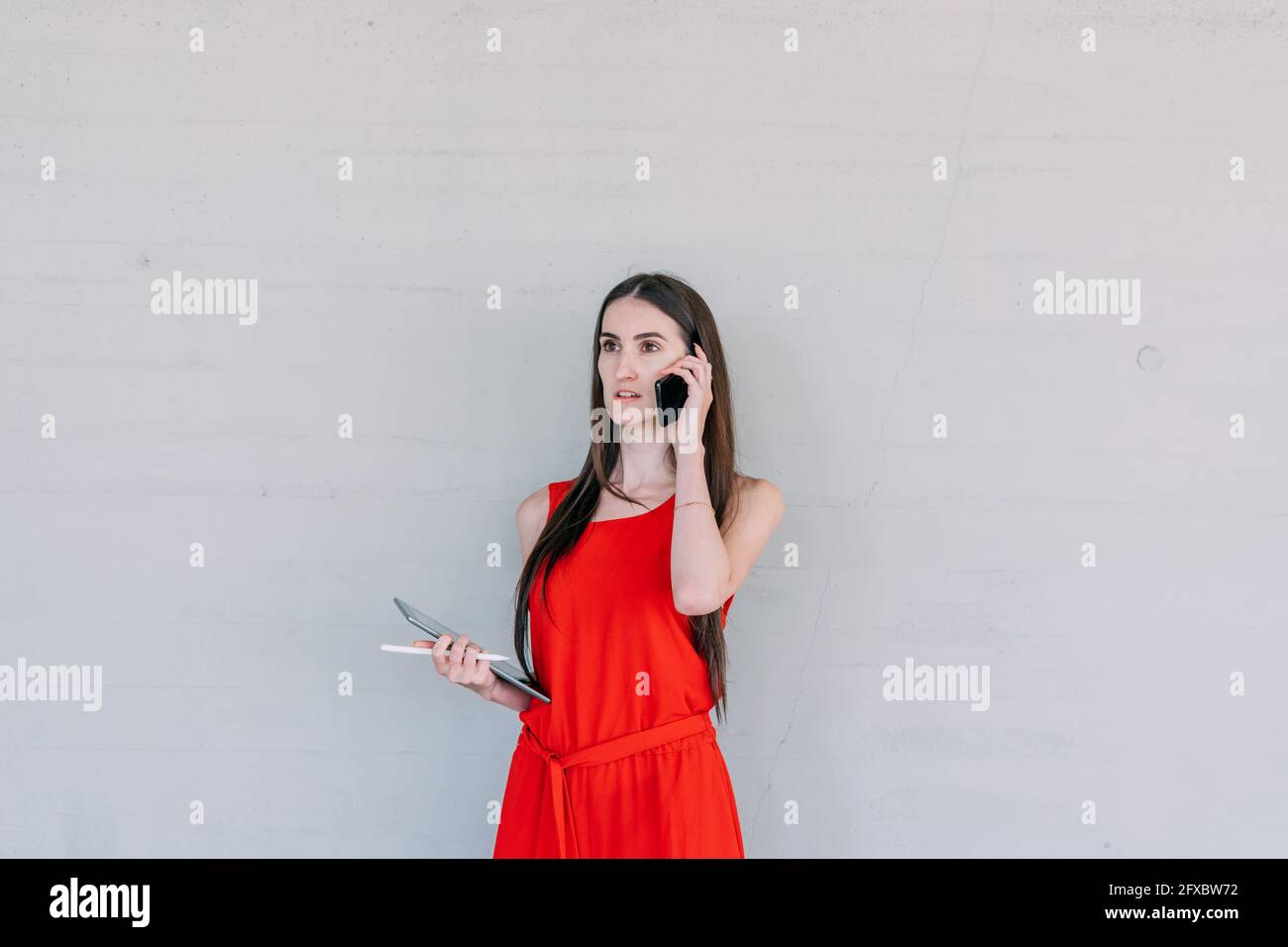 Young business woman in red dress, holding a tablet while receiving a phone call with her cell phone. Stock Photo