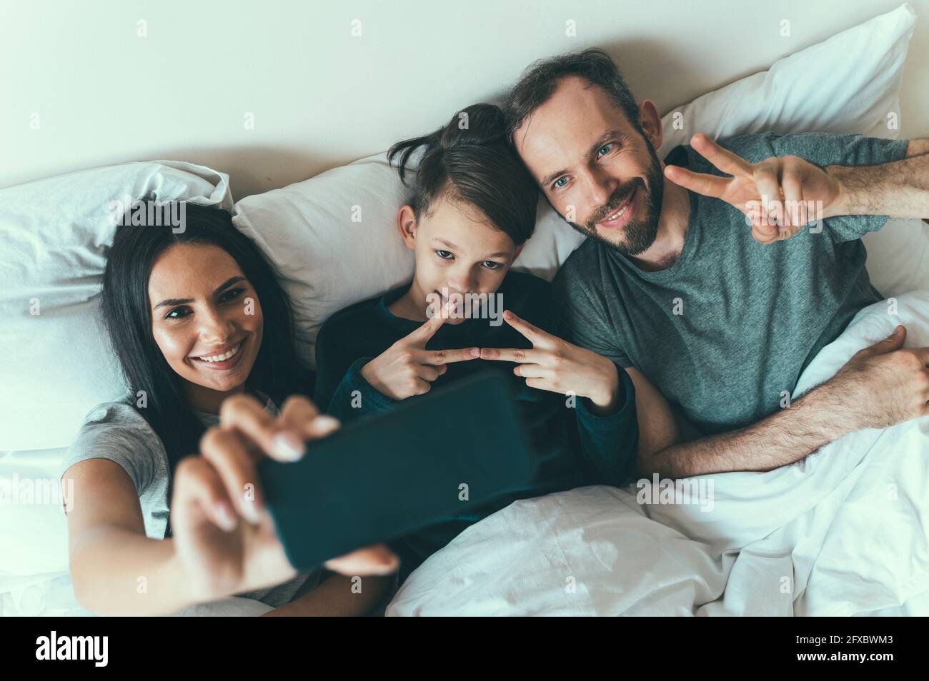 Father and son gesturing while woman taking selfie lying on bed at home Stock Photo