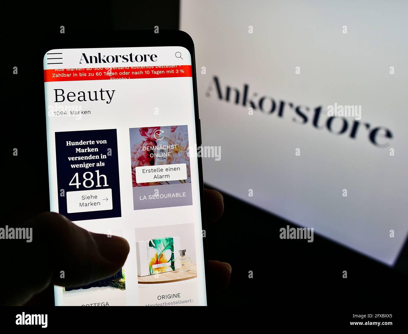 Person holding mobile phone with webpage of French B2B marketplace company Ankorstore on screen in front of logo. Focus on center of phone display. Stock Photo