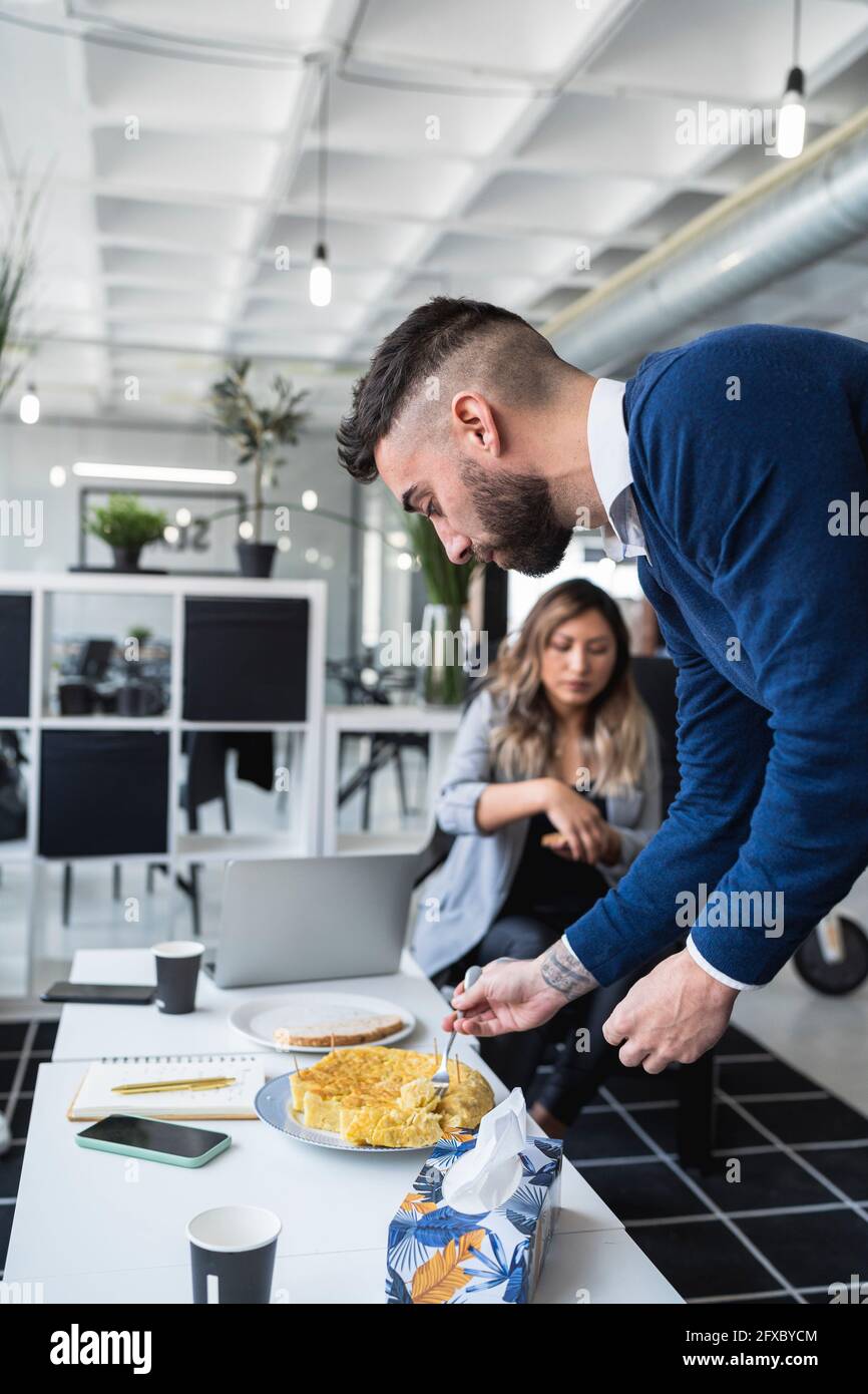 Businessman having Spanish omelet at table with female colleague in background at office Stock Photo