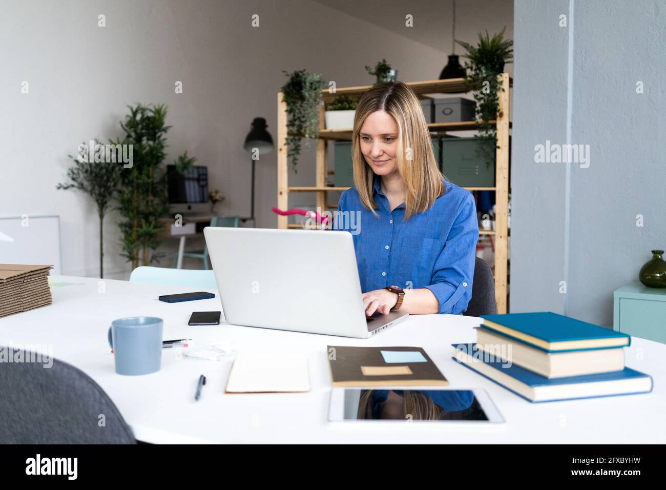 Smiling businesswoman with books on desk using laptop at office Stock Photo