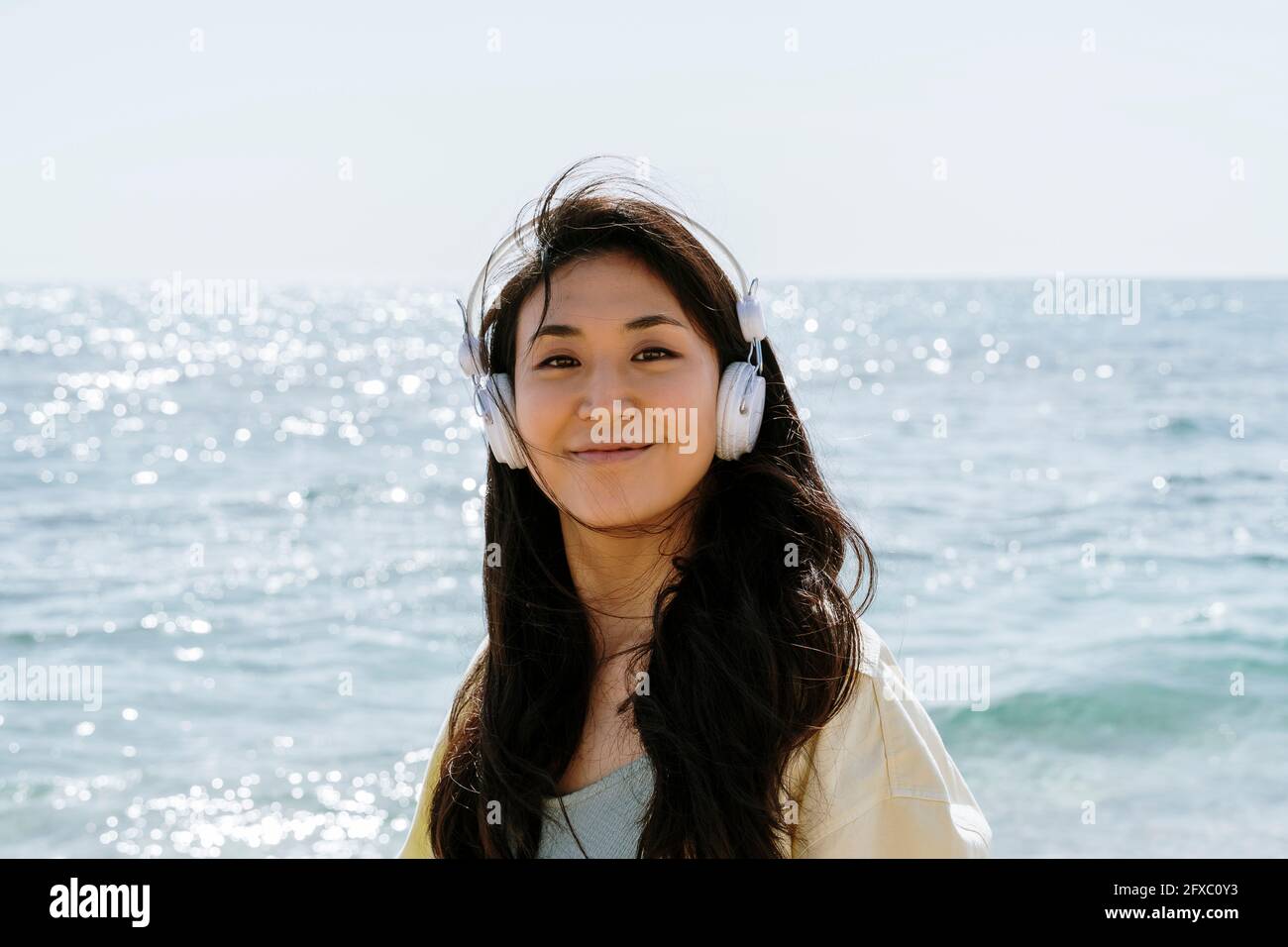Beautiful woman with in-ear headphones standing on beach during sunny day Stock Photo