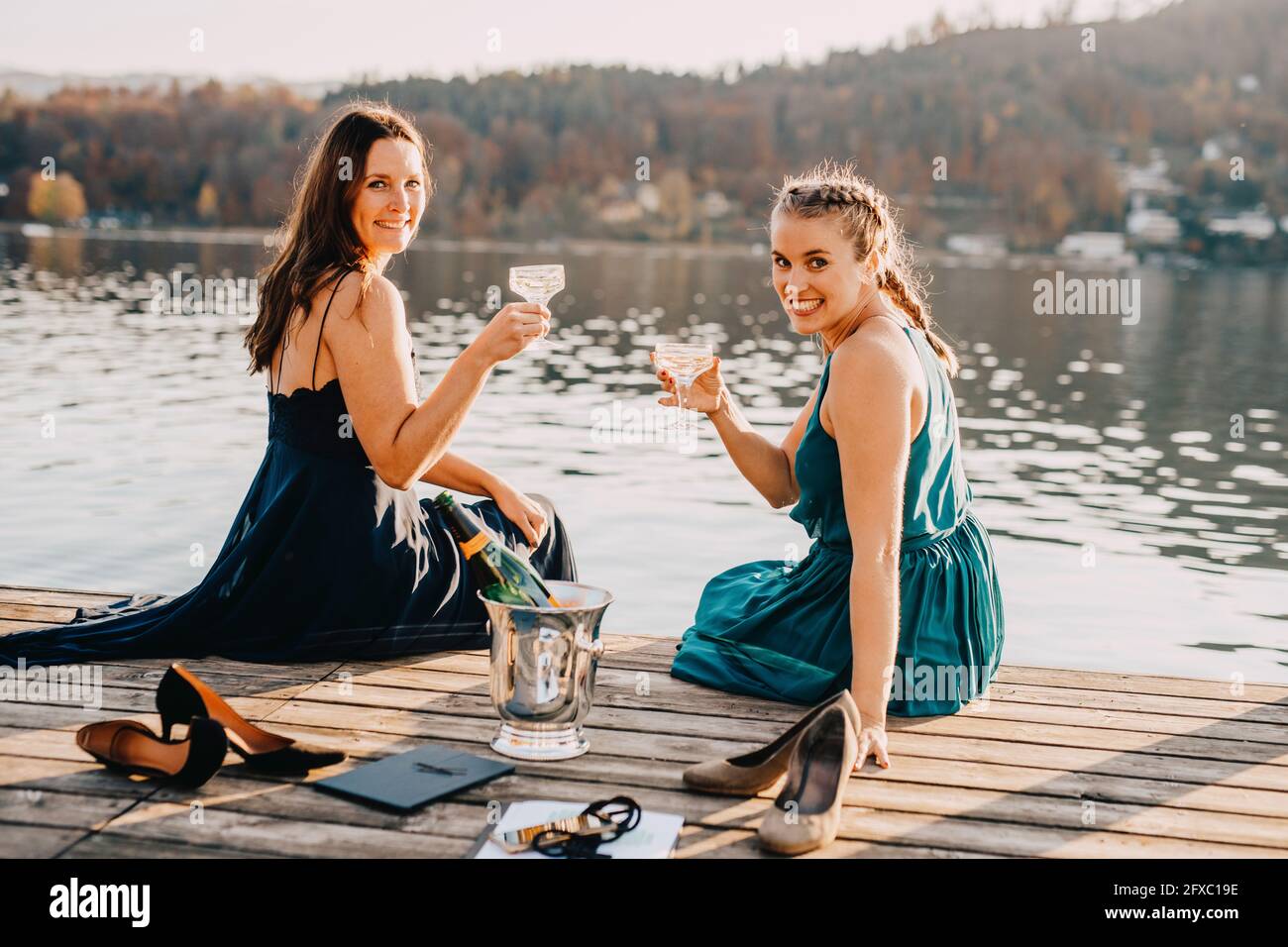 Smiling female event planners sitting with champagne on jetty over lake Stock Photo