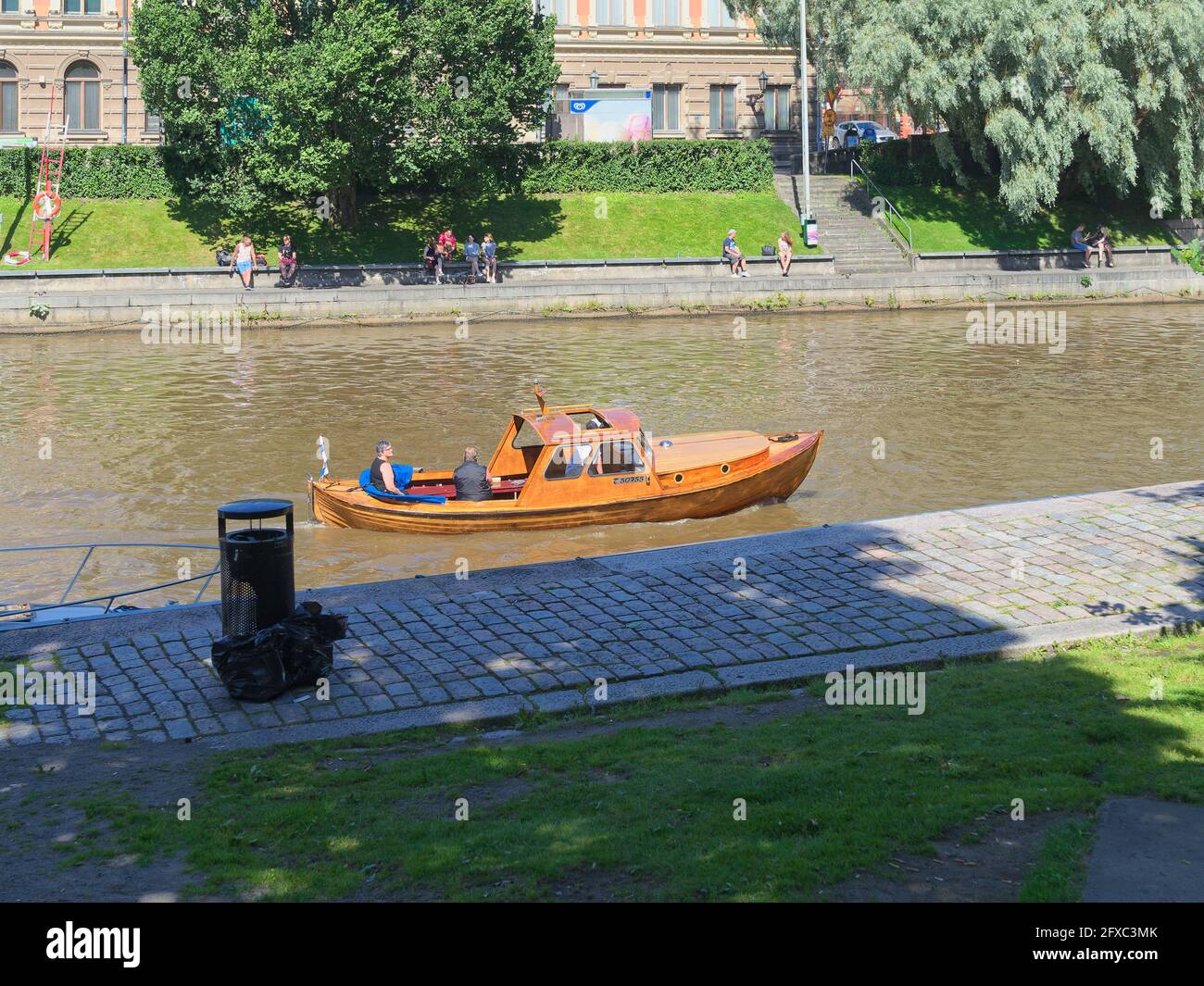 People enjoy a sunny summer day on the bank of river Aura in Turku, Finland while a wooden boat sails upstream. Stock Photo