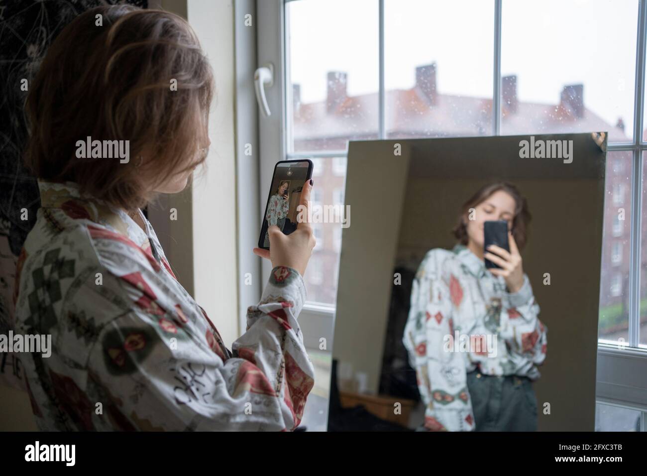 Young woman photographing reflection in mirror through smart phone at home Stock Photo