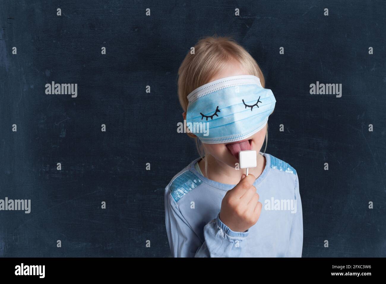 Girl with protective face mask licking lollipop against black background Stock Photo