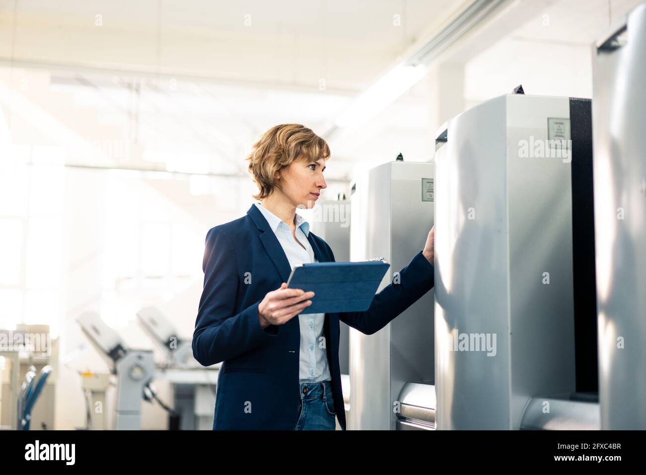 Female manager with digital tablet checking machine while standing in factory Stock Photo