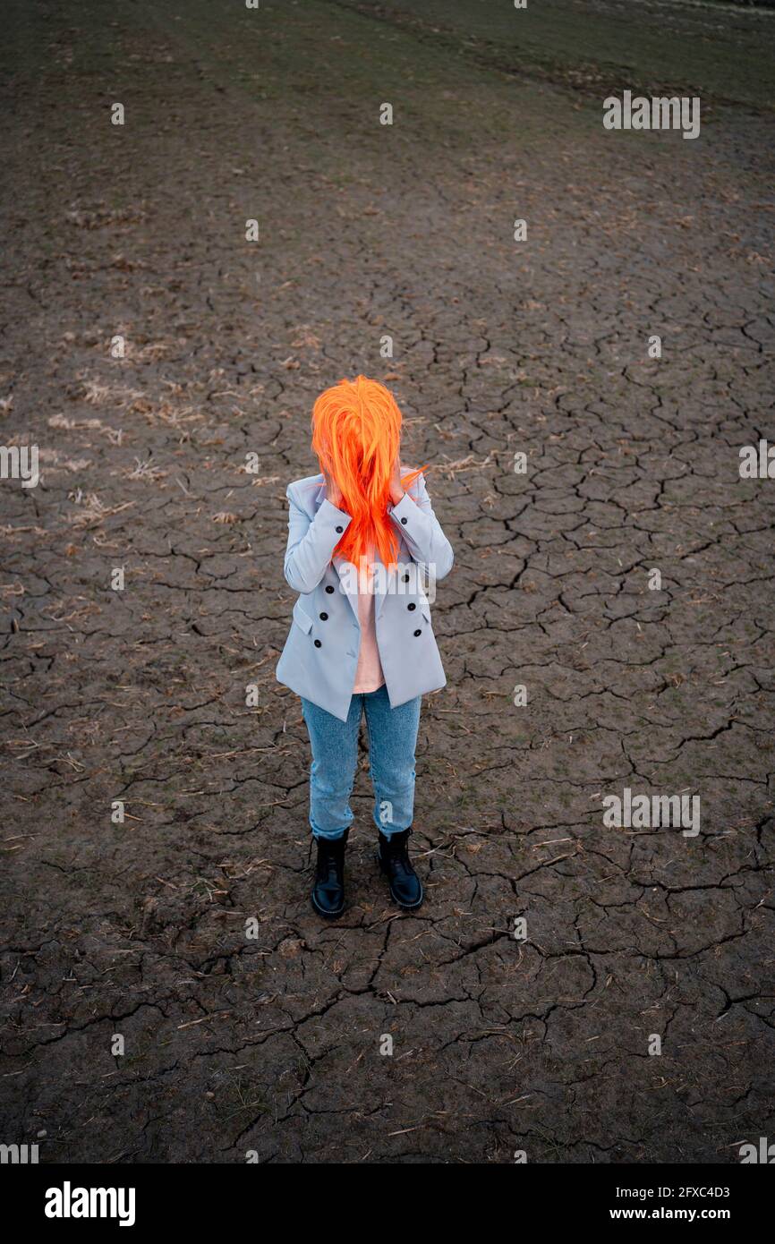 Young woman with face covered by orange hair wig on dirt field Stock Photo