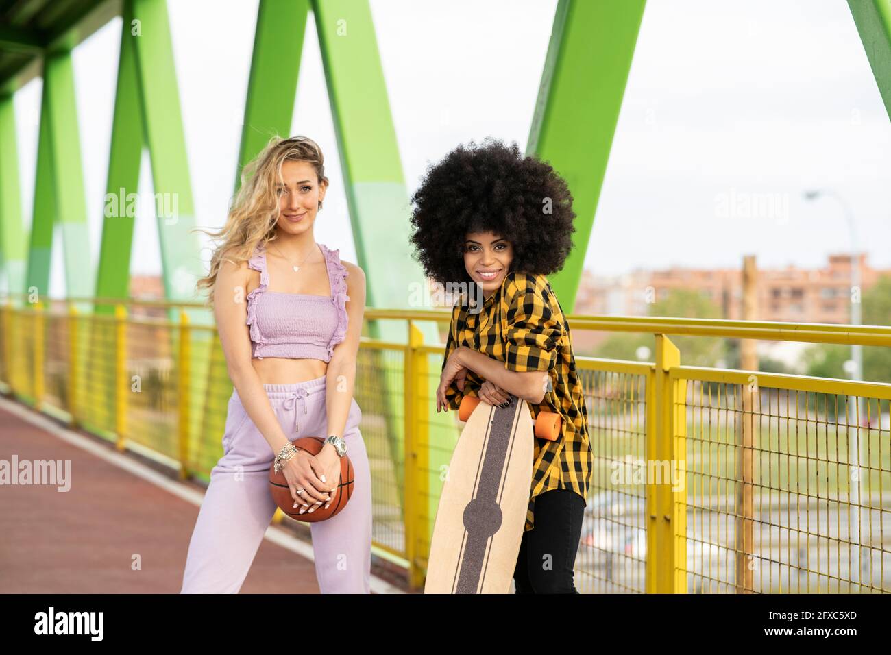 Multi-ethnic female friends with skateboard and basketball standing at bridge Stock Photo