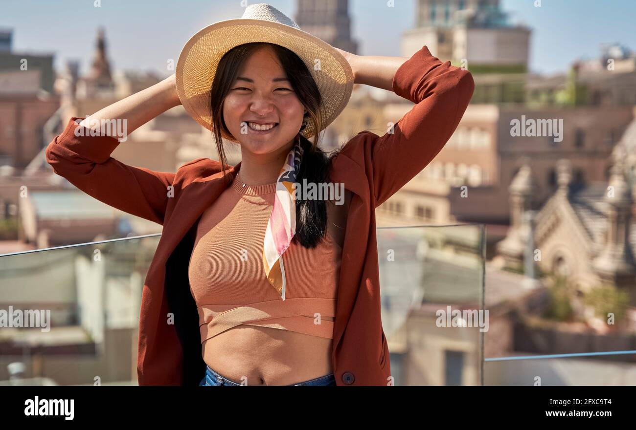 Smiling woman in cap standing with hands behind head at roof terrace Stock Photo