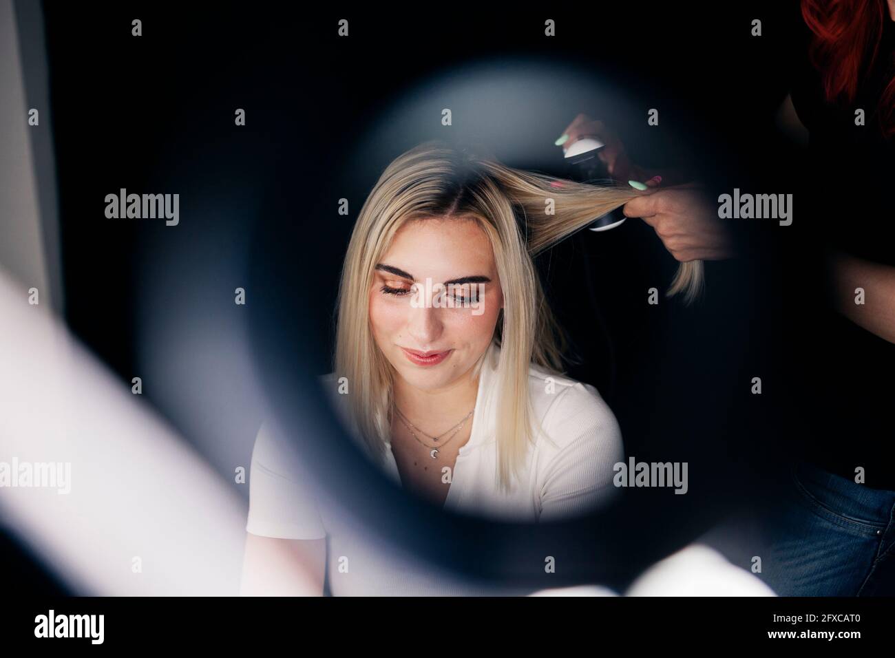 Female hairstylist using straightener on beautiful model's hair seen through ring light Stock Photo