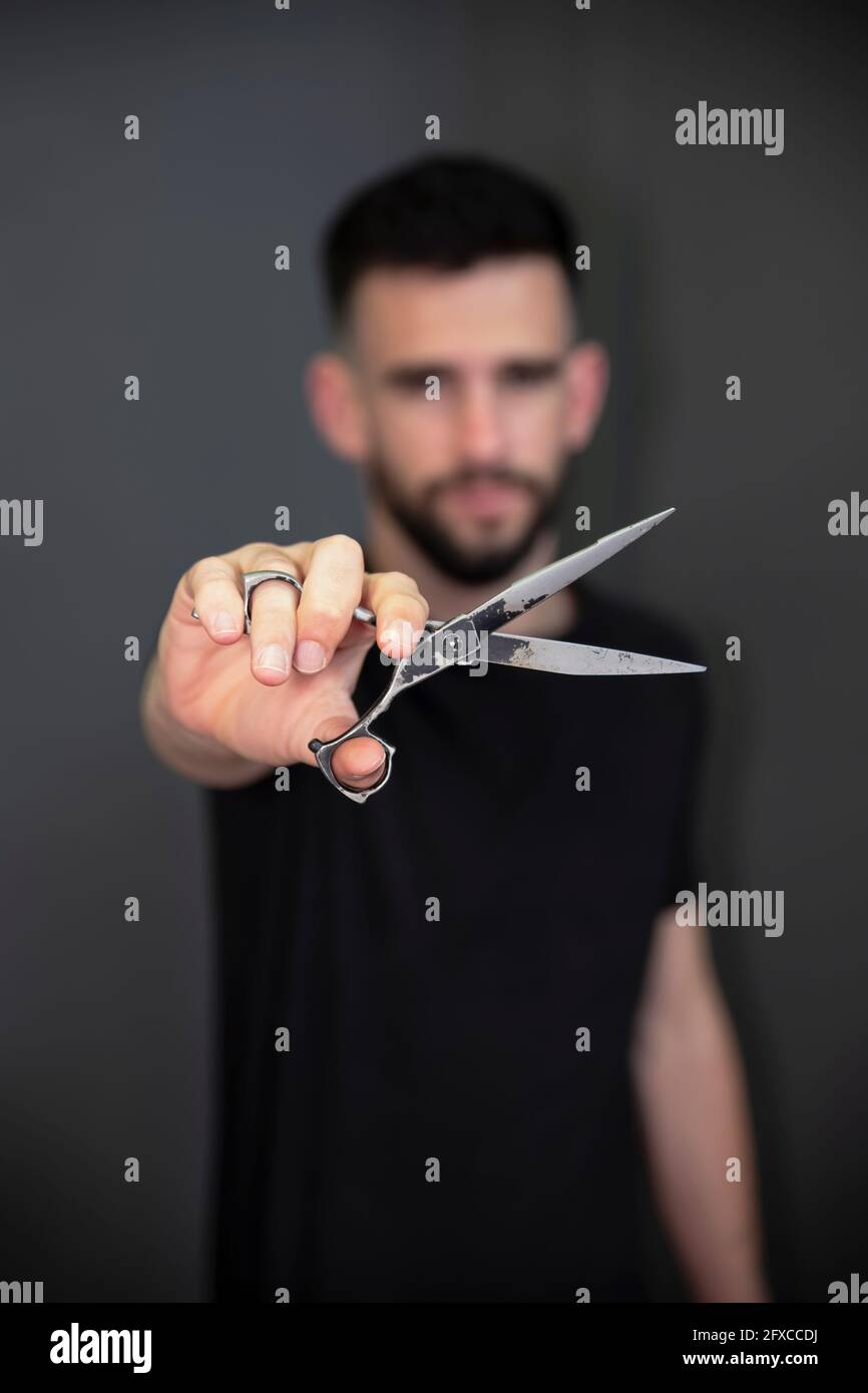 Young male barber showing scissor while standing in front of black wall at salon Stock Photo