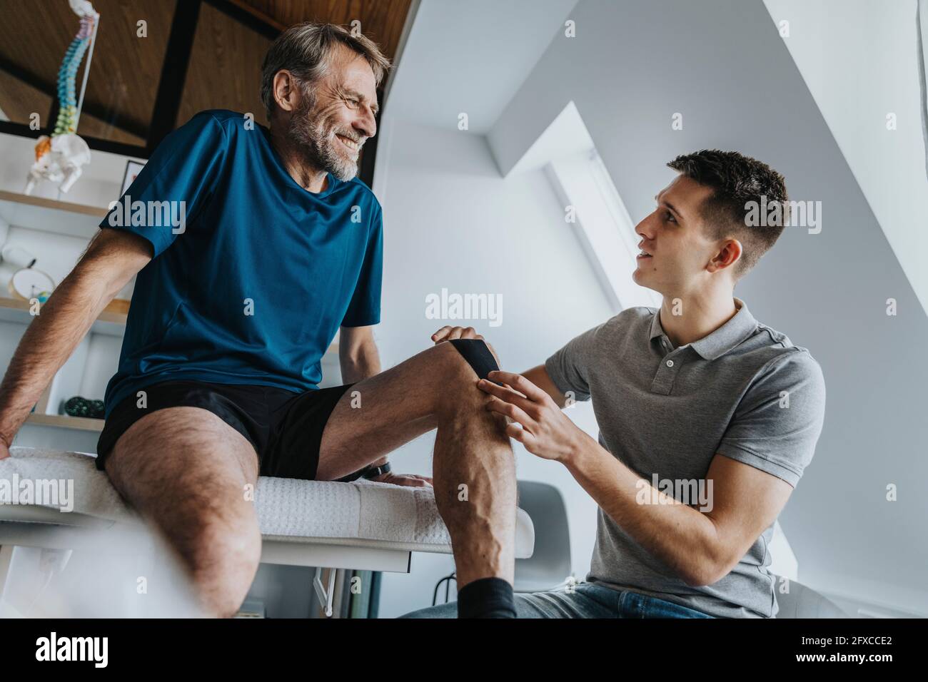 Male physical therapist sticking kinesio tape on smiling patient's knee in practice Stock Photo