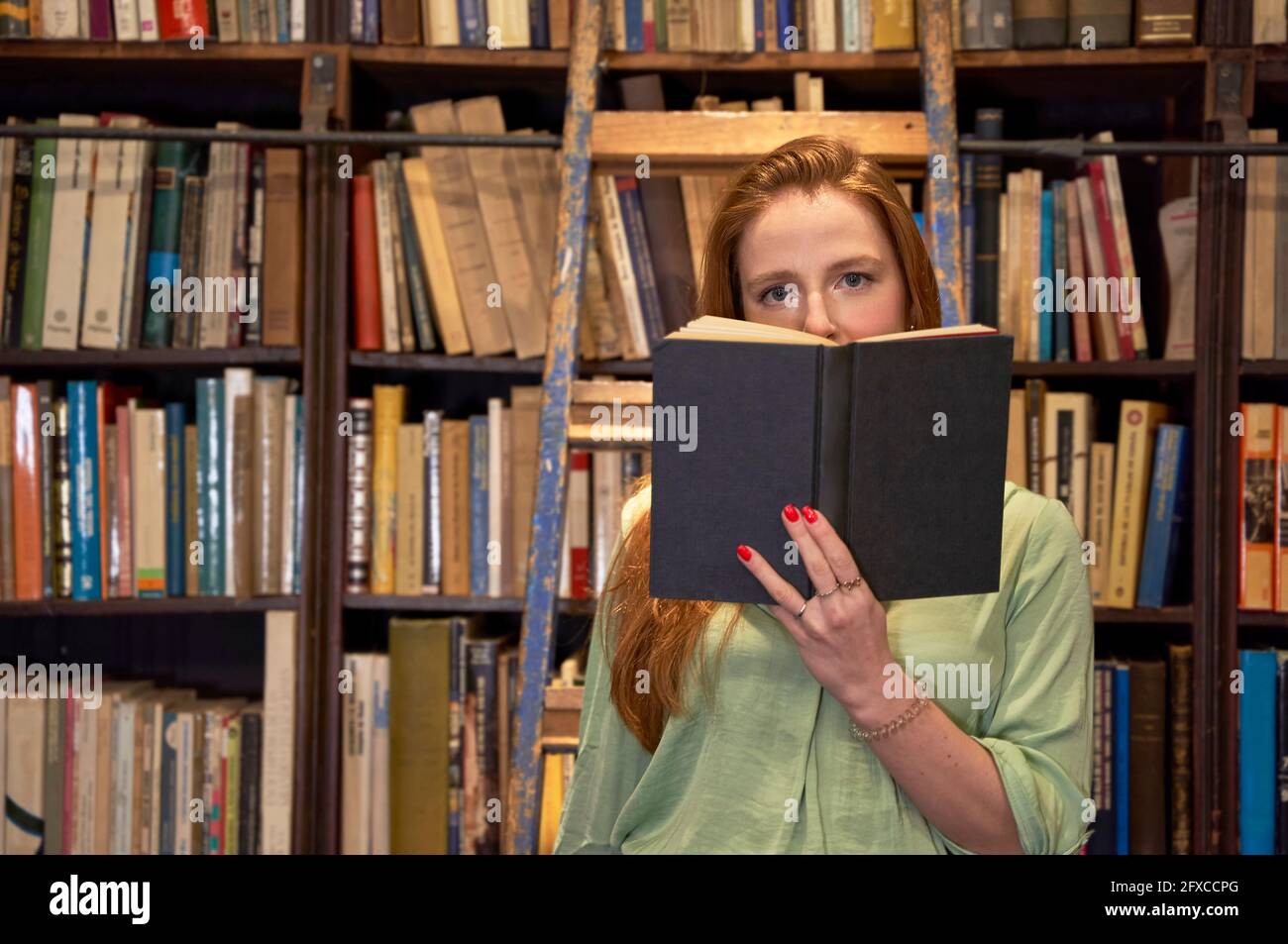 Young woman with book standing on ladder in library Stock Photo
