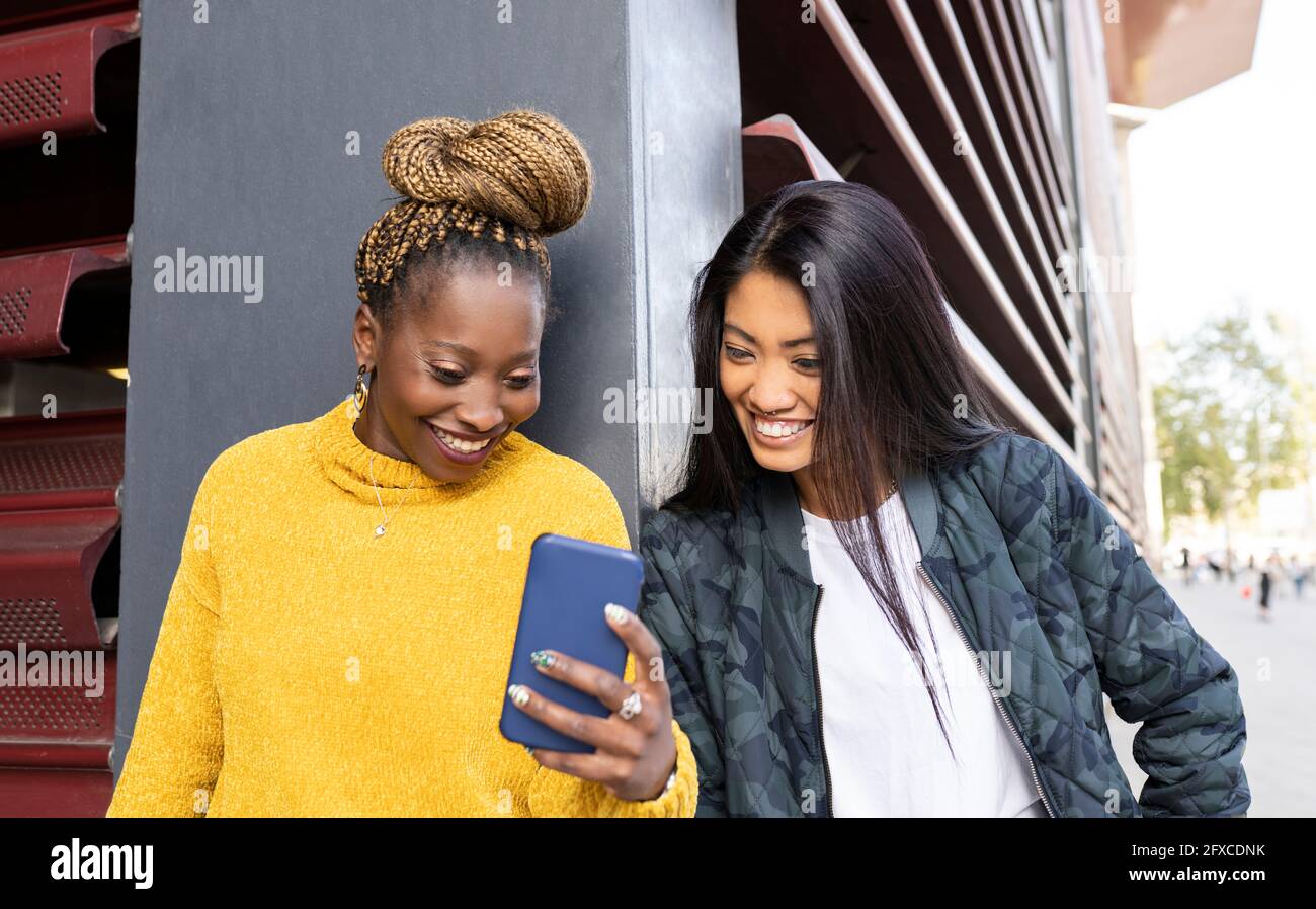Multi-ethnic female friends looking at smart phone by wall Stock Photo