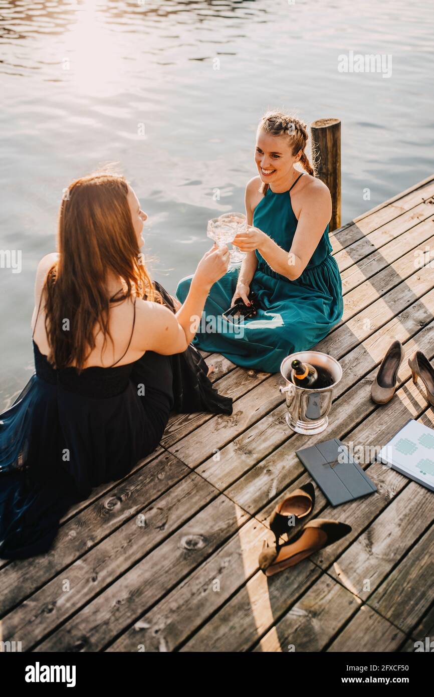 Smiling female event planners toasting champagne while sitting on jetty over lake Stock Photo