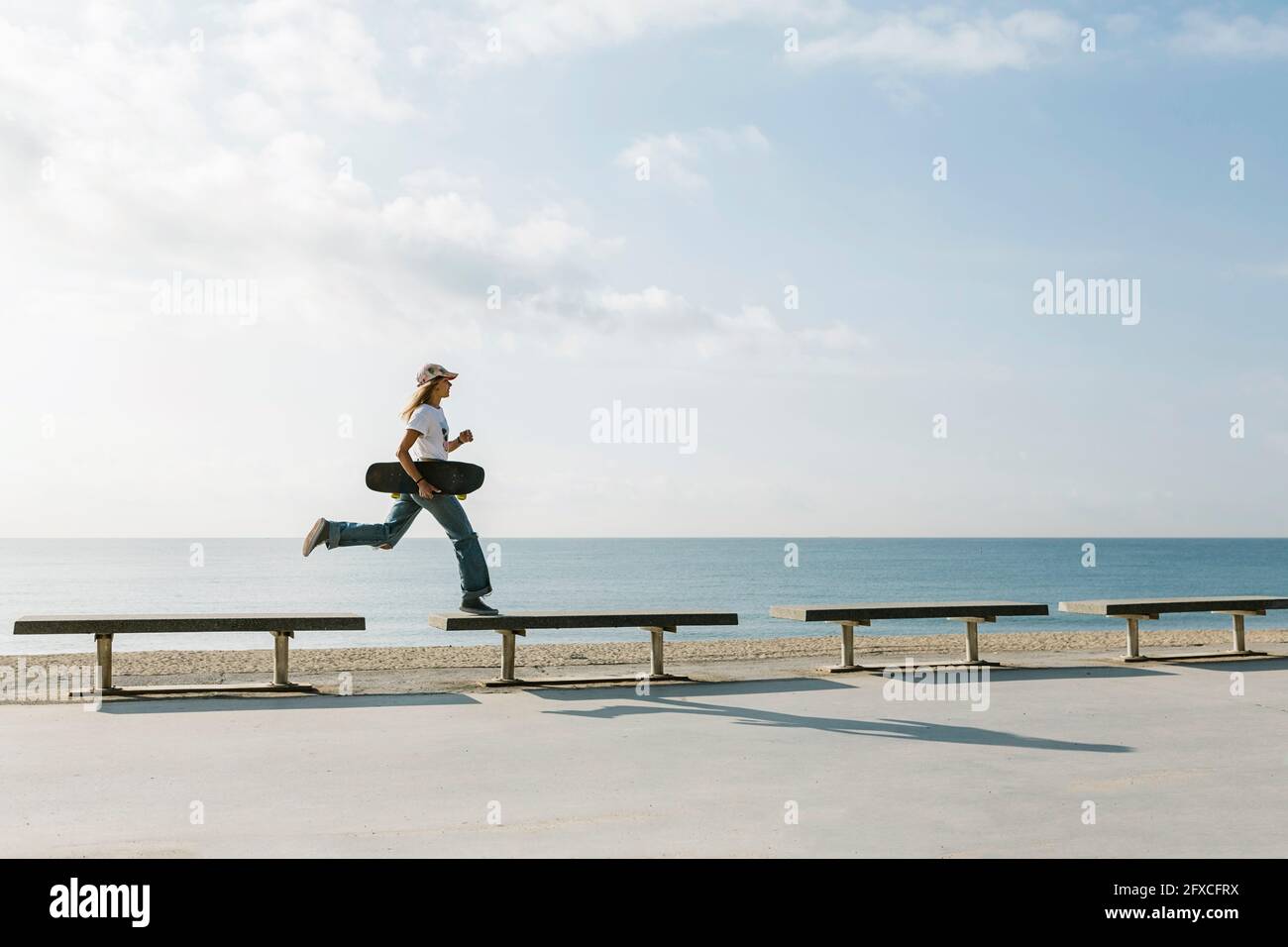 Woman with skateboard crossing bench while running at beach Stock Photo