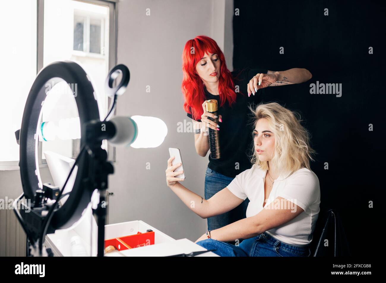 Female stylist applying spray on model's hair taking selfie through mobile phone in studio Stock Photo