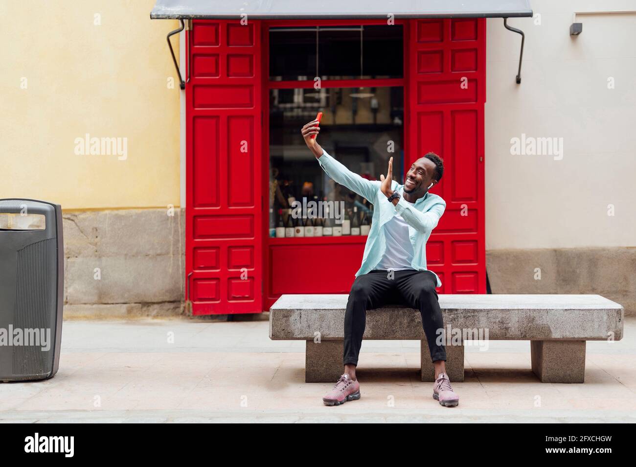 Young man waving during video call while sitting on bench outside store Stock Photo