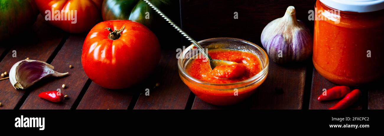 Homemade hot tomato sauce adjika in jars - Tomatoes, chilli pepper, garlic, herbs on wooden table. Stock Photo