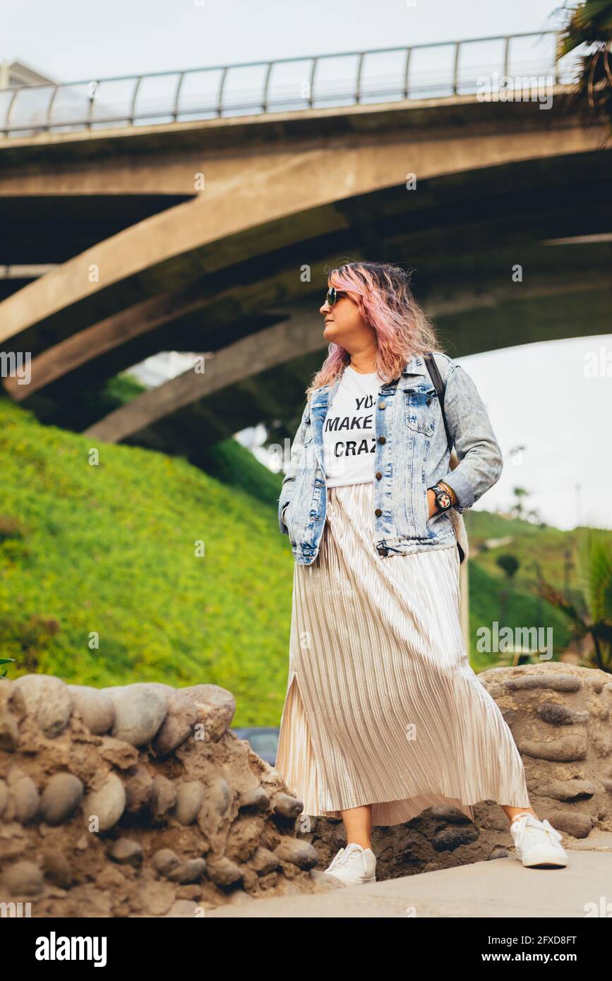 Full-length hipster woman standing with her hands on her jean coat, pink-painted hair and a silver-gold tone skirt outdoors, with a jumper behind. Ind Stock Photo