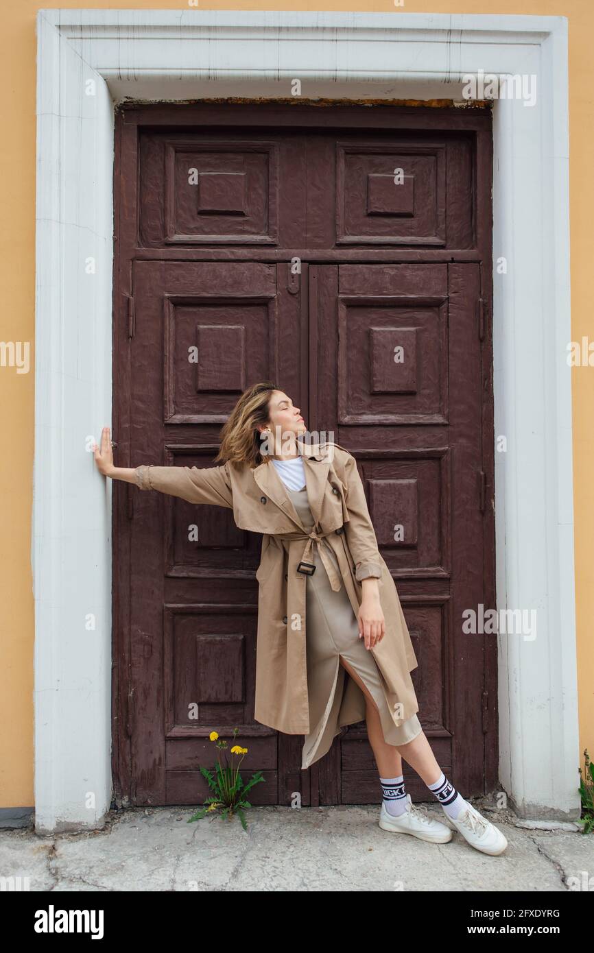 Young millennial woman with wild hair dressed in an autumn coat posing near the door of an old building. Stock Photo
