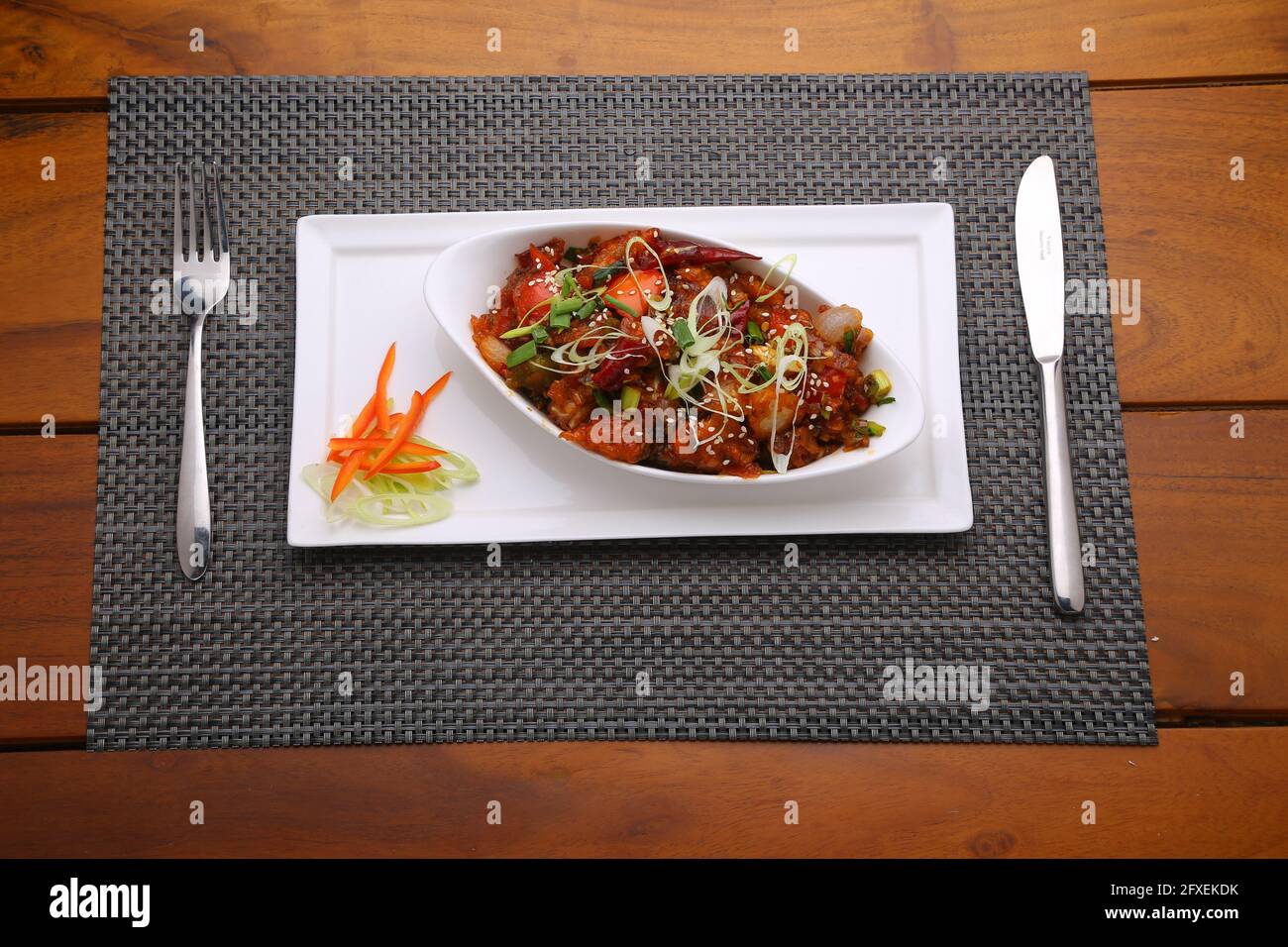 Chilli Chicken dry,well garnished and arranged in an oval shaped bowl on a white serving plate placed on a grey mat  with wooden texture Stock Photo
