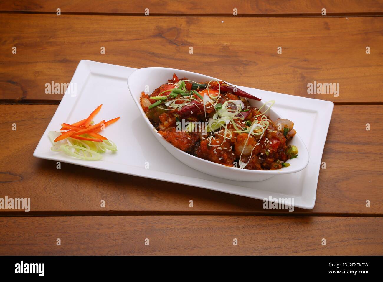 Chilli Chicken dry,well garnished and arranged in an oval shaped bowl on a white serving plate with wooden texture Stock Photo