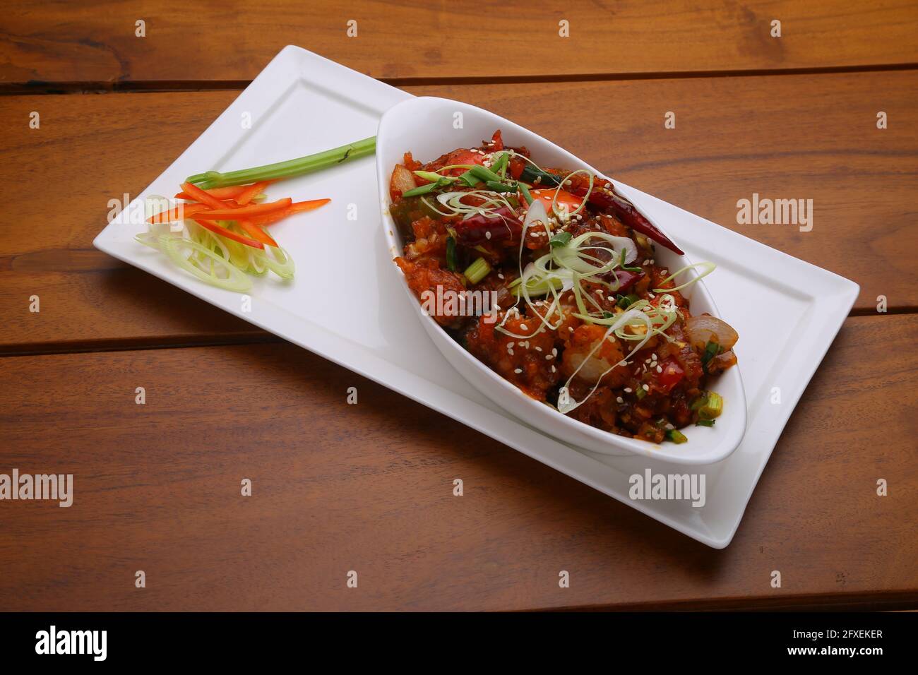 Chilli Chicken dry,well garnished and arranged in an oval shaped bowl on a white serving plate with wooden texture Stock Photo