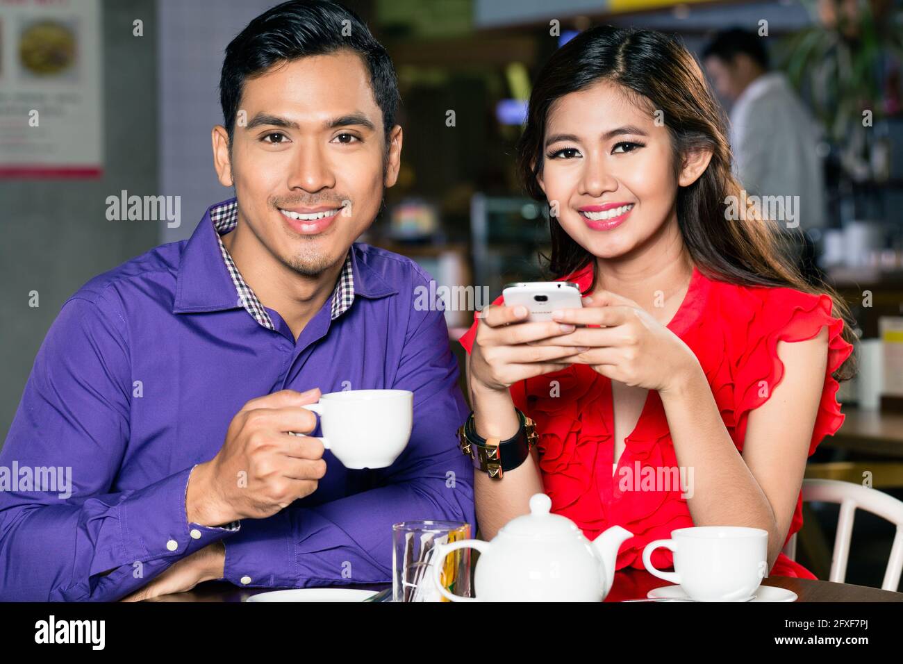 Portrait of a young and happy Asian couple looking at camera while relaxing together at table in a trendy coffee shop Stock Photo