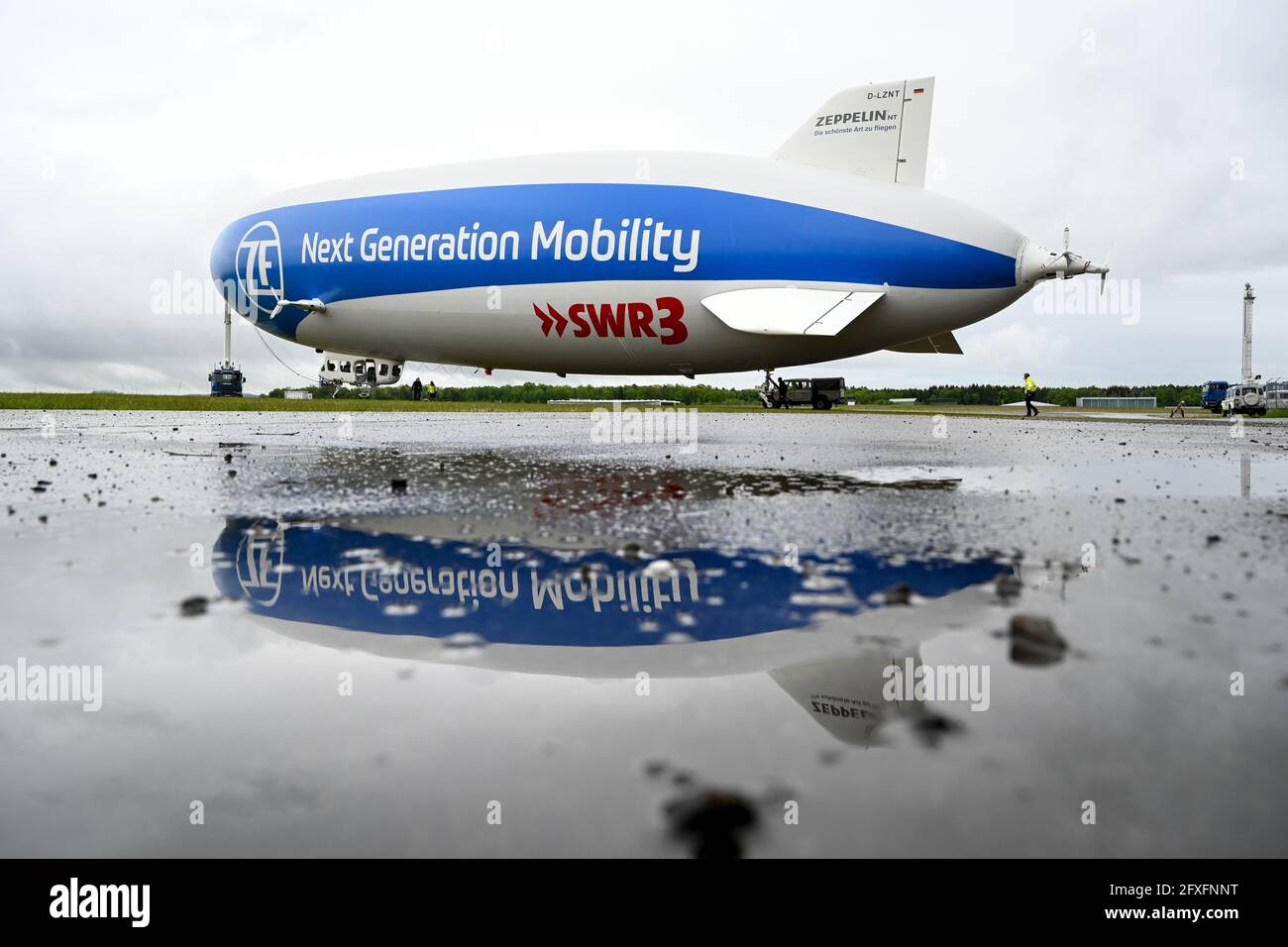 Friedrichshafen, Germany. 27th May, 2021. The Zeppelin NT (New Technology) is pulled out of the hangar by a truck so that the airship can take off for its press flight in the afternoon. The Zeppelin itself is reflected in a puddle in front of the hangar. The Zeppelin NT will start its season on Friday, 28.05.2021. Credit: Felix Kästle/dpa/Alamy Live News Stock Photo