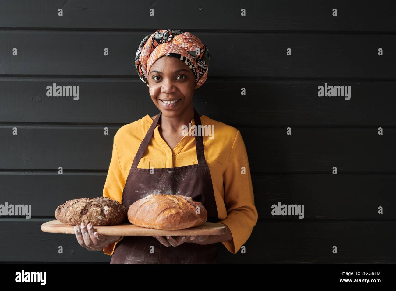 Portrait of young baker in apron holding the tray with fresh bread and smiling at camera Stock Photo