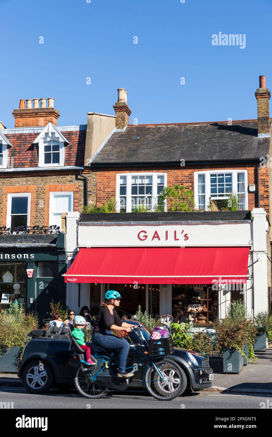 A young woman transports her child by bicycle past Gail's bakery in Dulwich Village, London, England, UK. Stock Photo