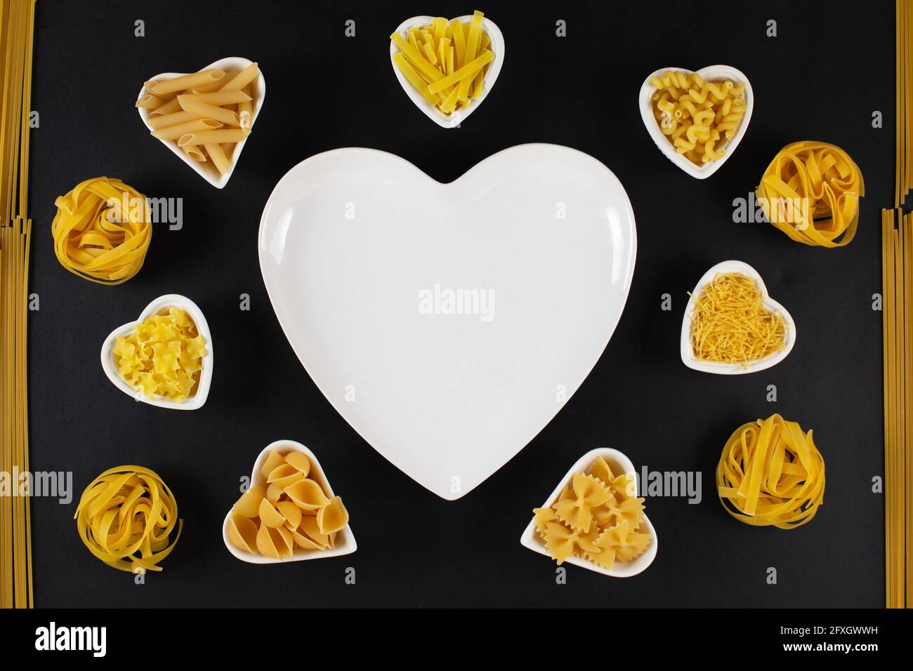 Pasta food selection around heart shaped porcelain dishes over black background. Uncooked, Dried pasta noodles. Love pasta concept Stock Photo
