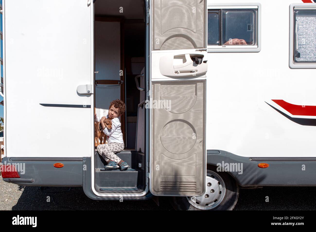 little curly-haired boy smiling sitting in the door of motor home, tenderly holding his pet. Stock Photo