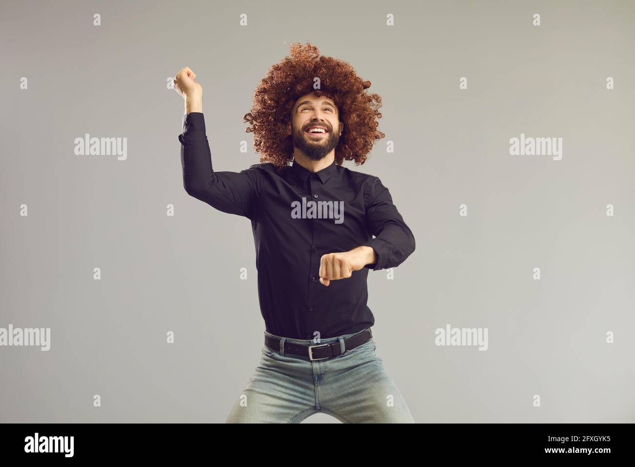 Happy young man in funny curly wig dancing gangnam style on gray studio background Stock Photo