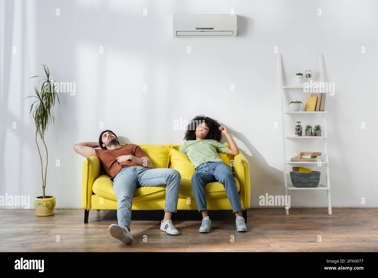 exhausted multiethnic couple sitting on yellow sofa and suffering from heat in summer Stock Photo