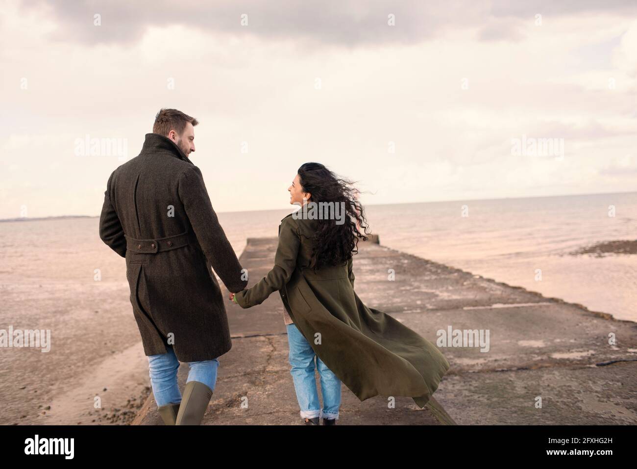 Couple in winter coats holding hands on ocean beach jetty Stock Photo